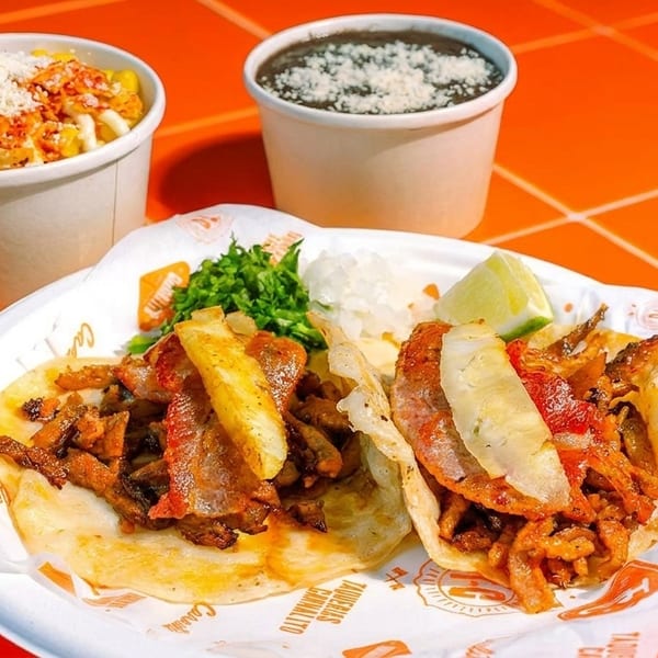 Two tacos with meat, pineapple, cilantro, and onions, with sides of elote and black beans.