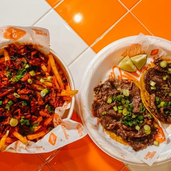 Loaded fries and two carne asada tacos with lime wedges on an orange and white tiled surface.