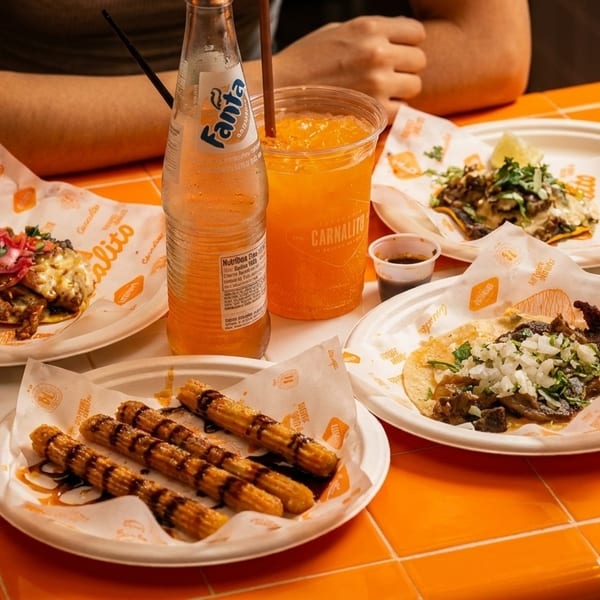 A vibrant orange tiled table laden with tacos, churros, Fanta, and Modelo Chelada.